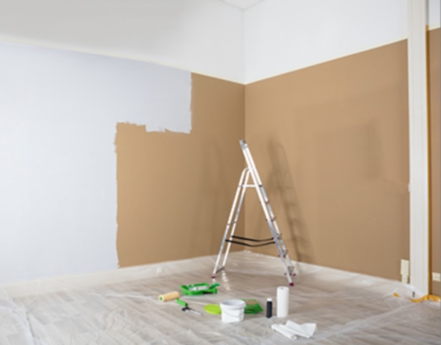Pokój w trakcie malowania: ściana częściowo biała, częściowo brązowa, drabina, wałek i kuweta malarska. Podłoga zabezpieczona folią malarską.