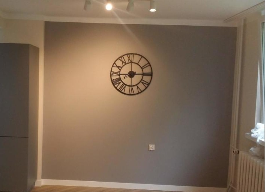 Ściana pomalowana na szaro z dekoracyjnym zegarem w stylu vintage, listwa przypodłogowa biała, oświetlenie punktowe na suficie, widoczny fragment szafki.