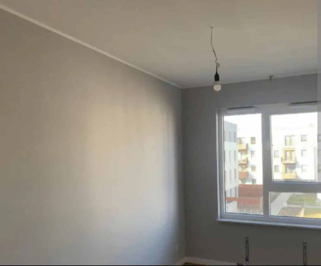 Świeżo pomalowane szare ściany w pokoju z oknem i wiszącą żarówką. Widok na budynek z balkonami za oknem. Listwa przysufitowa.