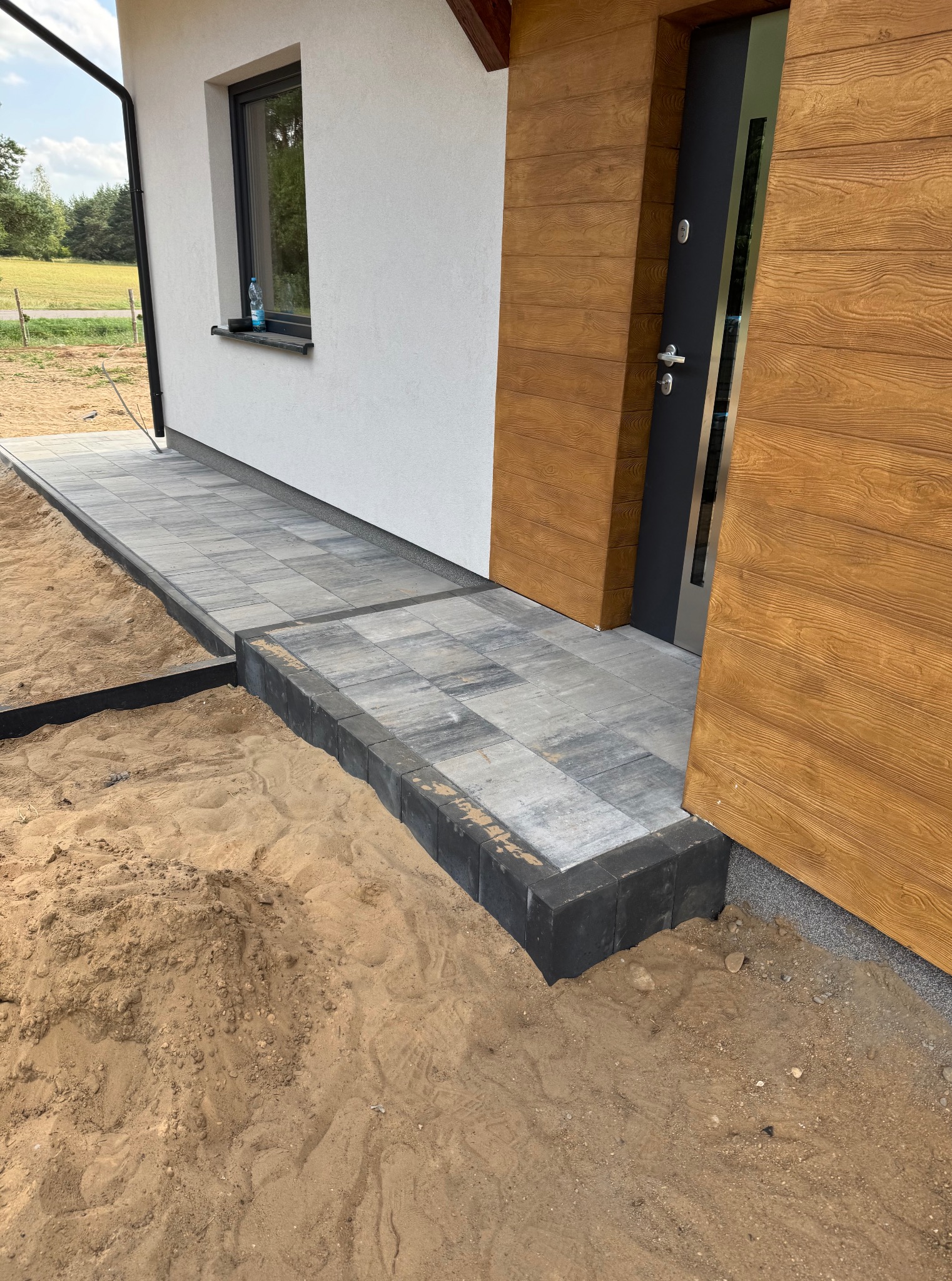 Wykończony chodnik z kostki brukowej prowadzący do drzwi wejściowych domu. Widoczny piasek i obrzeża. Detal z bliska.