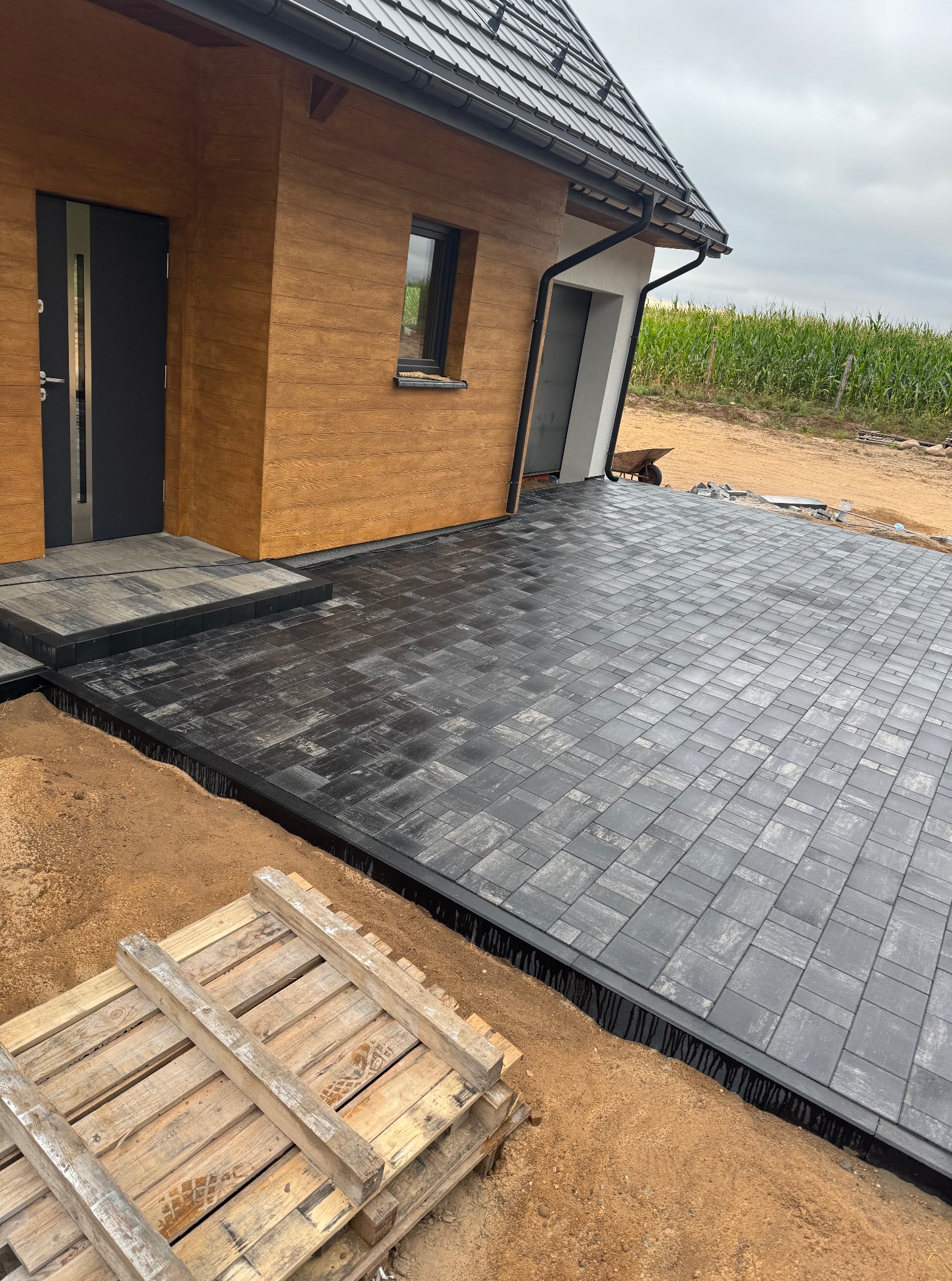 Nowoczesny podjazd z szarej kostki brukowej przed domem z drewnianą elewacją. Widoczny paleta i przygotowany teren pod dalsze prace.