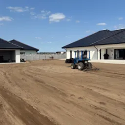 Niwelacja terenu pod ogród: mały traktor wyrównuje ziemię na działce przed nowoczesnym domem z czarnym dachem i garażem. W tle widoczny fragment ogrodzenia.