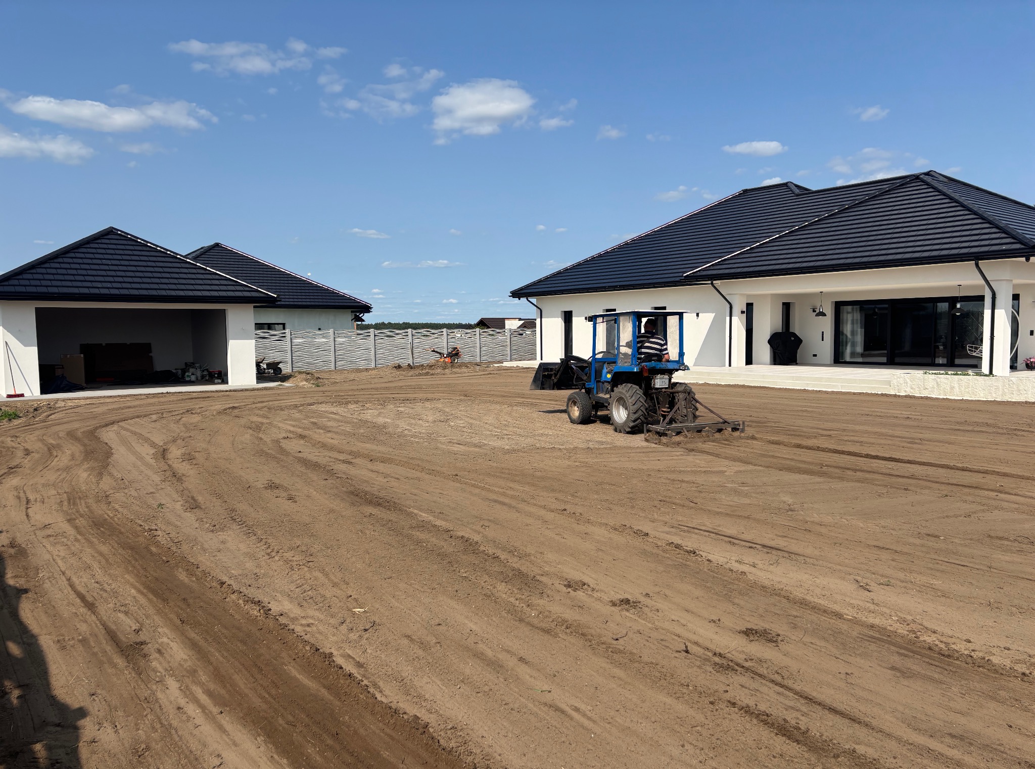 Niwelacja terenu pod ogród: mały traktor wyrównuje ziemię na działce przed nowoczesnym domem z czarnym dachem i garażem. W tle widoczny fragment ogrodzenia.