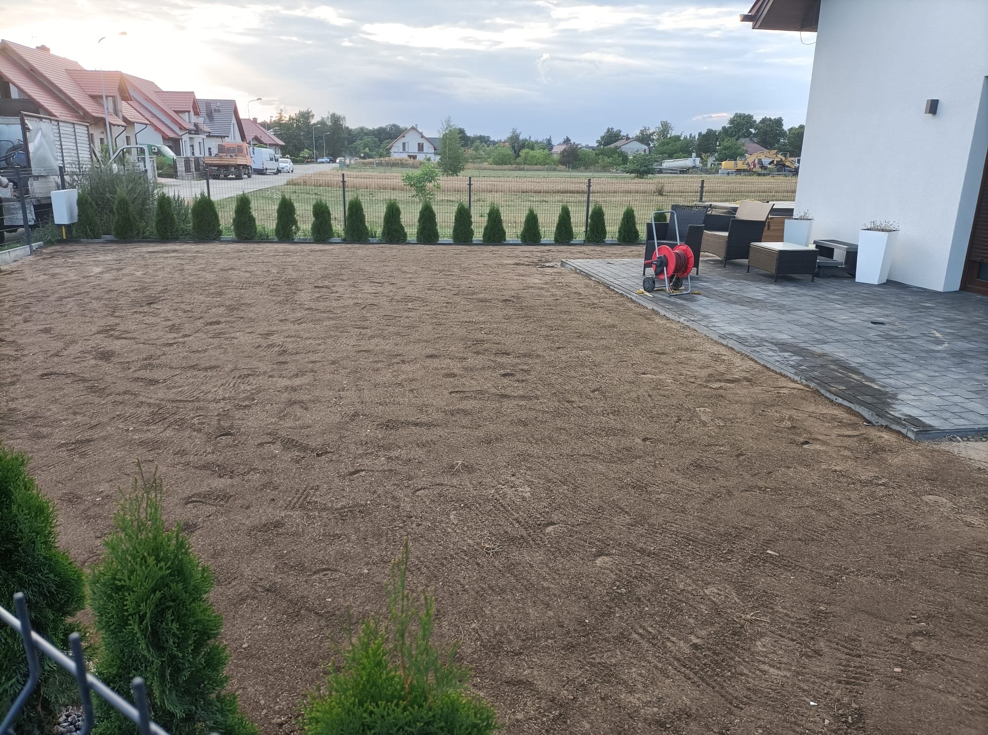 Przygotowany teren pod trawnik, równa powierzchnia ziemi w ogrodzie. W tle rząd posadzonych drzewek iglastych, obok taras z meblami ogrodowymi.