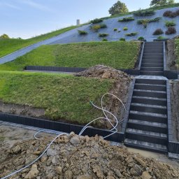 MAT-BRUK - Budowa schodów z kostki brukowej na wzgórzu z roślinami, widoczny etap prac, krawężniki i stos ziemi, biały wąż do podlewania. Nietypowe ujęcie w budownictwie.