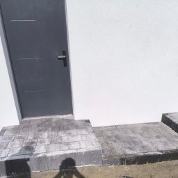 MAT-BRUK - Schody wejściowe z kostki brukowej w odcieniach szarości, prowadzące do szarych drzwi na tle białej ściany budynku. Widoczny fragment powierzchni z kostki.