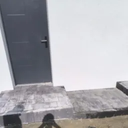 Schody wejściowe z kostki brukowej w odcieniach szarości, prowadzące do szarych drzwi na tle białej ściany budynku. Widoczny fragment powierzchni z kostki.