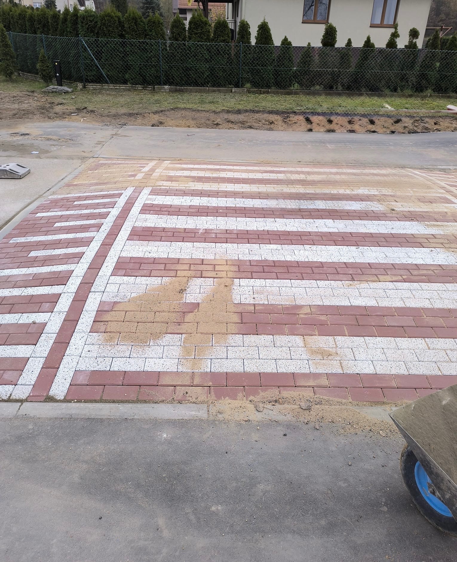 Częściowo ułożona kostka brukowa w biało-czerwone pasy z dekoracyjnym wzorem. Widoczny wózek budowlany i piasek do fugowania. Tło: trawnik i dom.