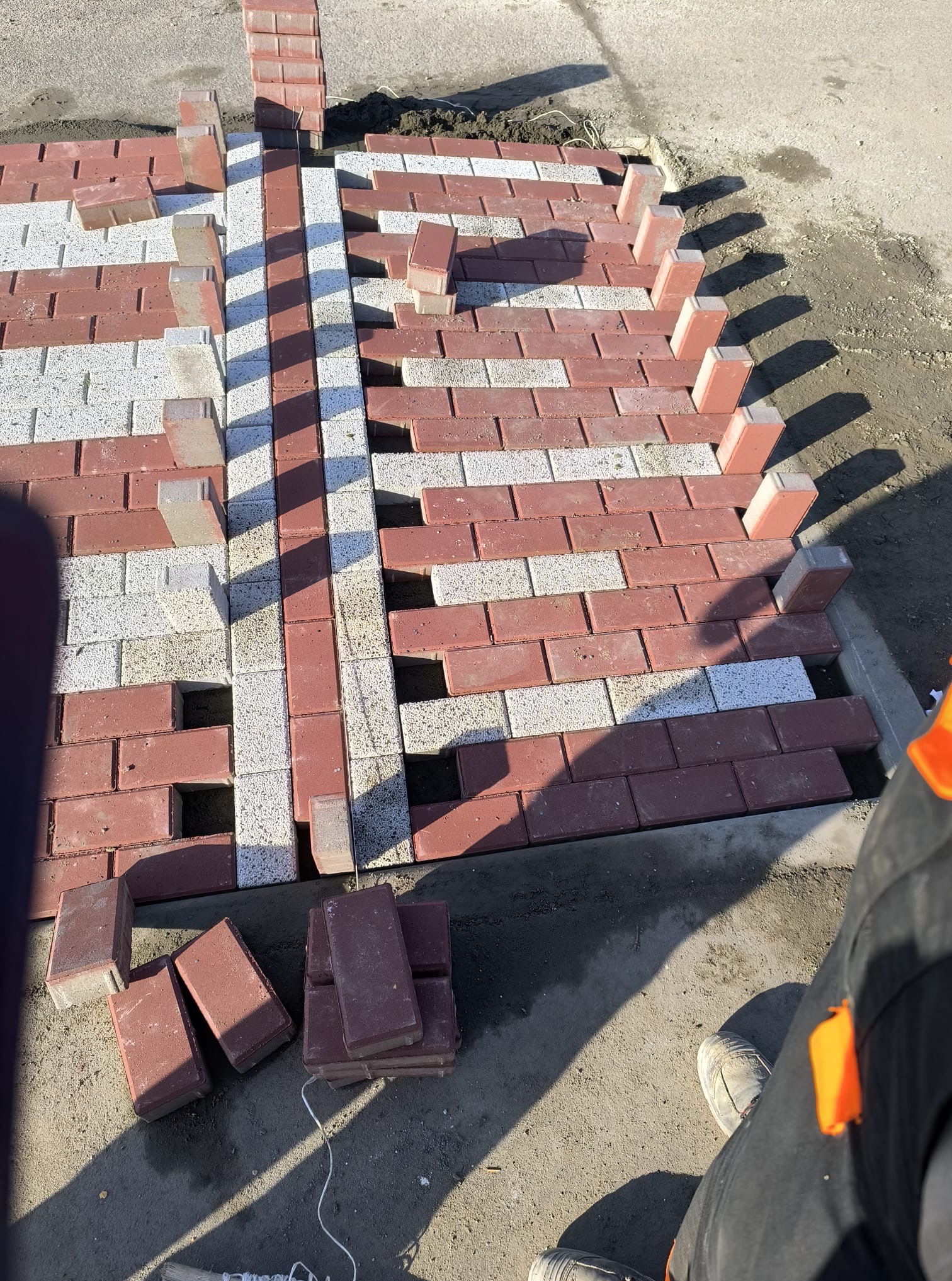 Fragment układania dwukolorowej kostki brukowej w geometryczny wzór. Widoczne niedokończone rzędy i zapas kostki. Praca w toku, w słoneczny dzień.