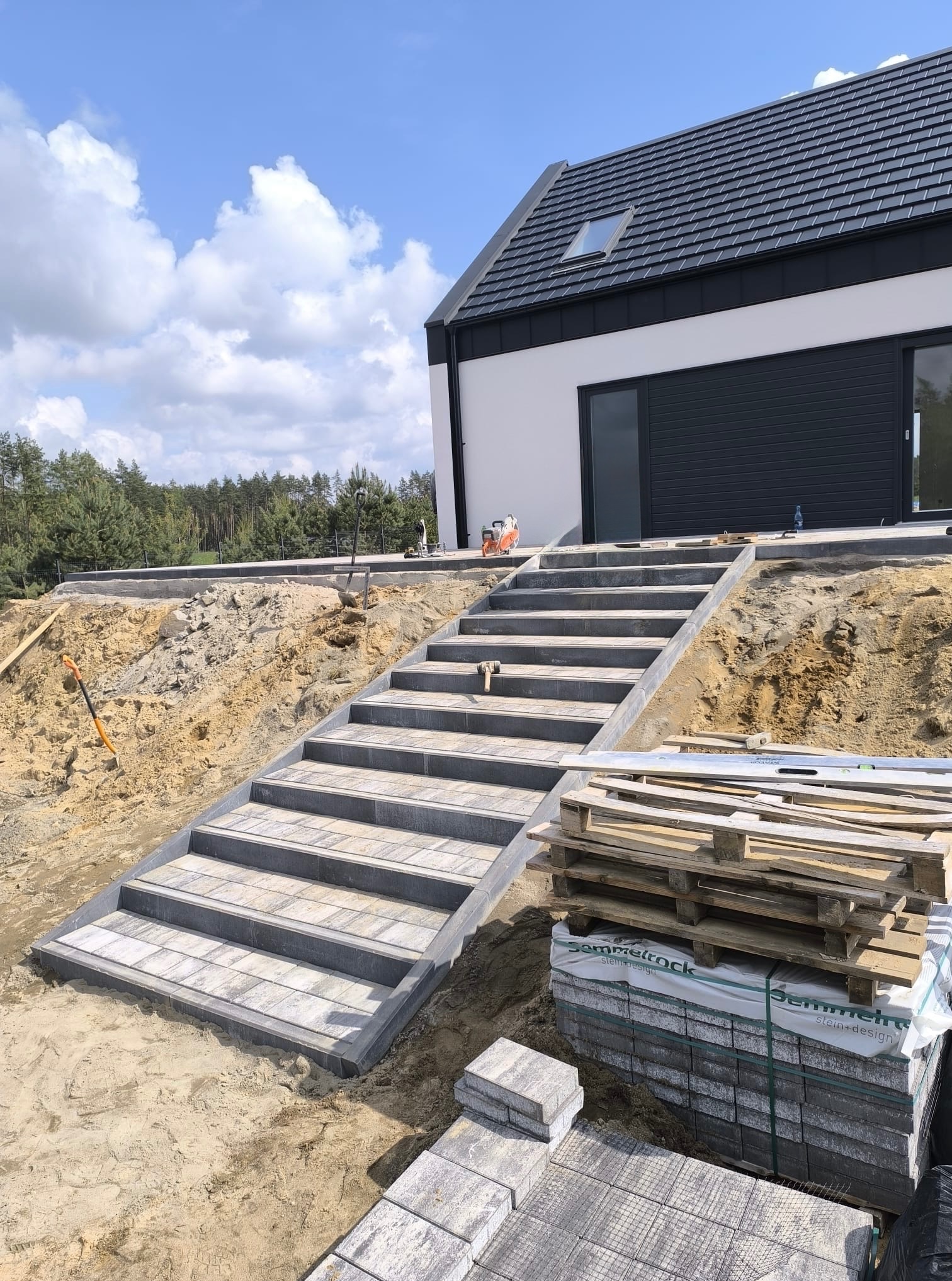 Schody z kostki brukowej prowadzące do nowoczesnego domu z szarą stolarką. Paleta z kostką brukowa w prawym dolnym rogu. Etap prac w toku.