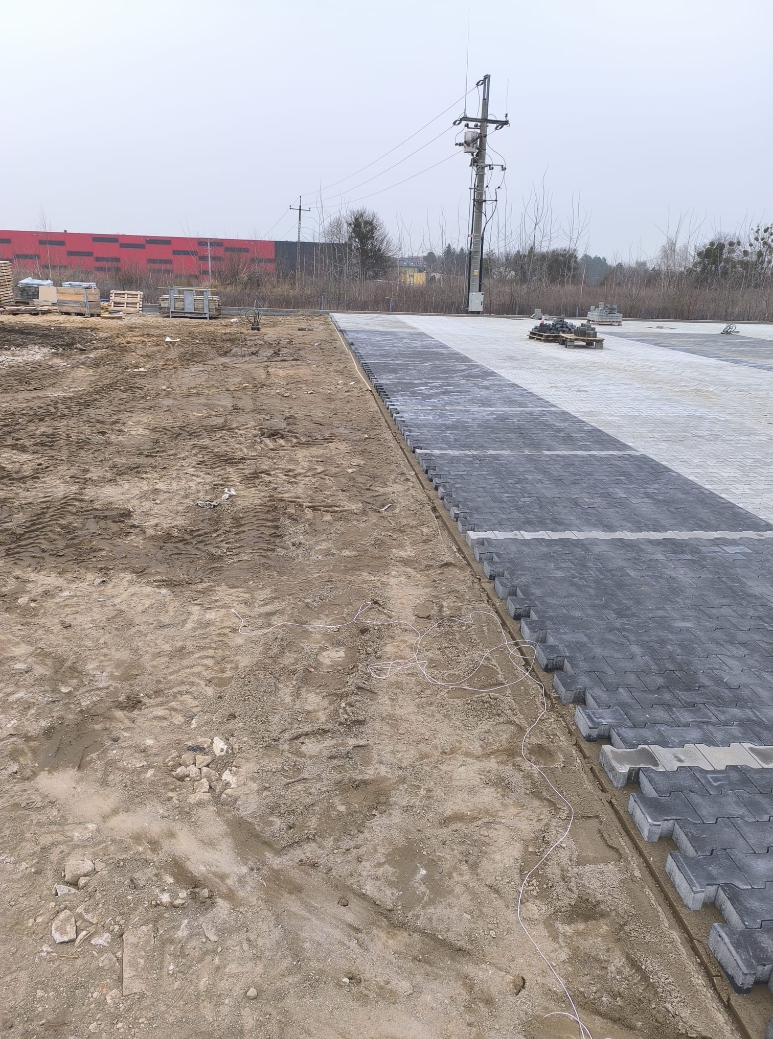 Fragment układanej nawierzchni z szarej kostki, widoczne etapy prac, ziemia i ułożona część. W tle budynek i słup elektryczny.