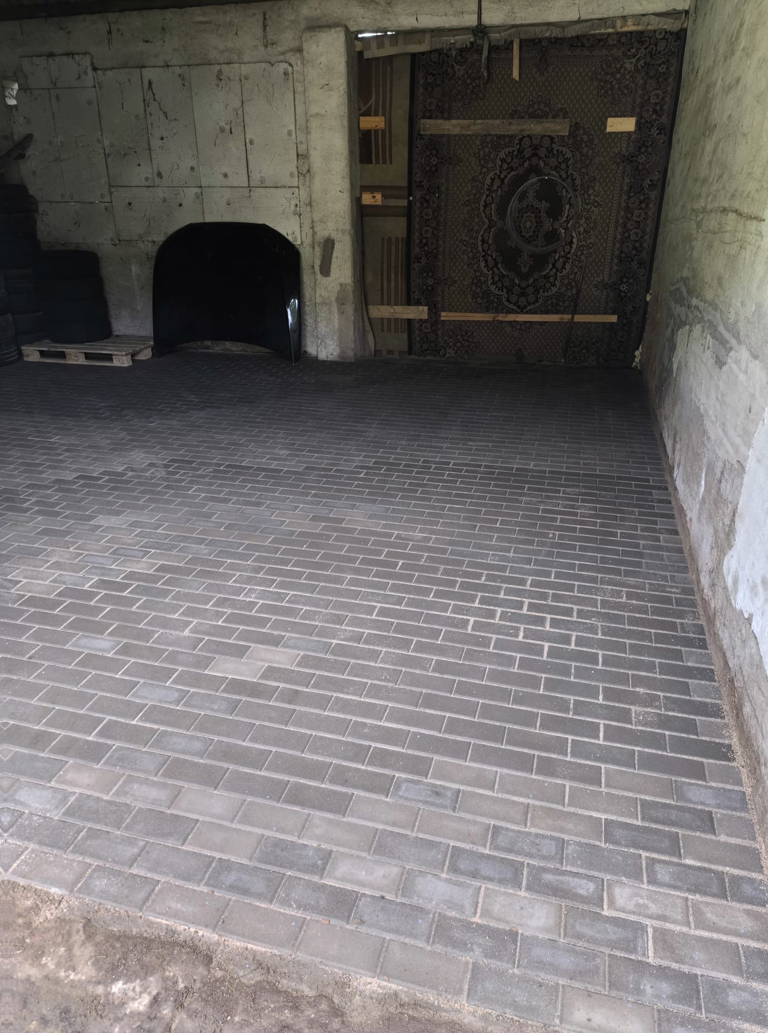 Ułożona szara kostka brukowa w garażu w Lęborku. Widoczna część palety z kostką. Nietypowe tło z dywanem na ścianie i elementami konstrukcyjnymi.