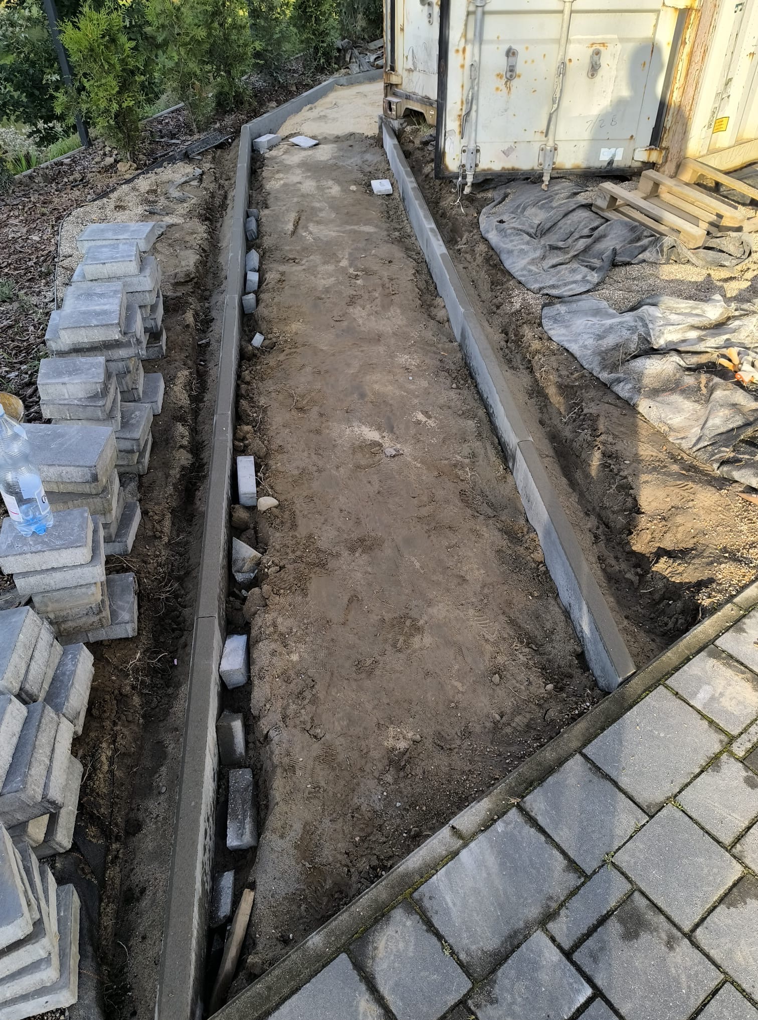 Przygotowanie podłoża pod ścieżkę z kostki brukowej. Widoczne obrzeża, podsypka i stosy kostki. Etap prac przed ułożeniem nawierzchni.