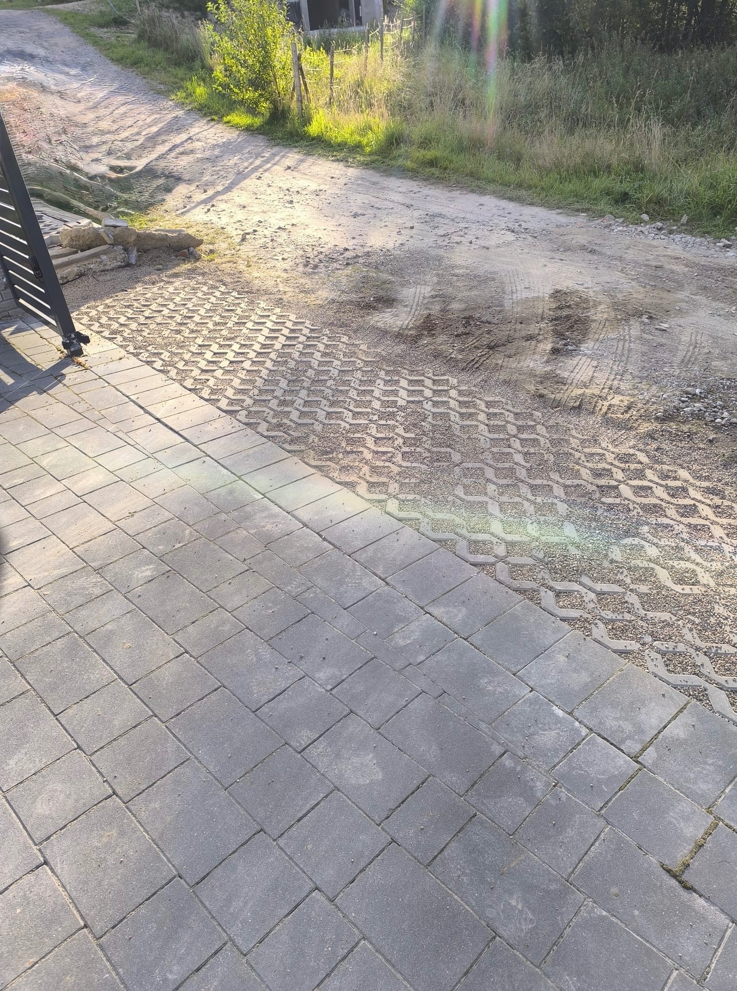 Fragment posesji z ułożoną szarą kostką brukową i ażurową, wypełnioną żwirem. W tle droga gruntowa i zieleń. Widoczny narożnik metalowej bramy.