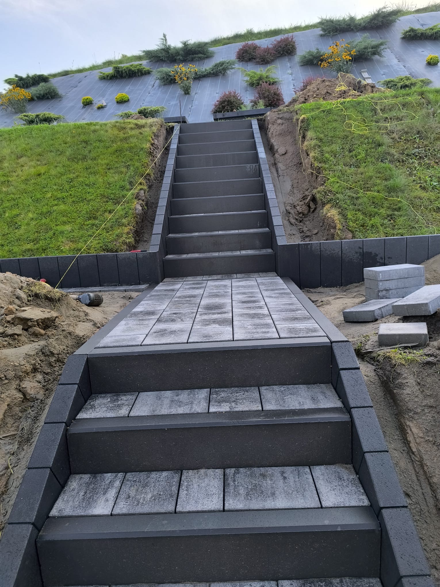 Schody z kostki brukowej prowadzące na wzgórze z roślinnością, obrzeża z ciemnej kostki, częściowo ułożone, widoczne narzędzia i materiały budowlane.