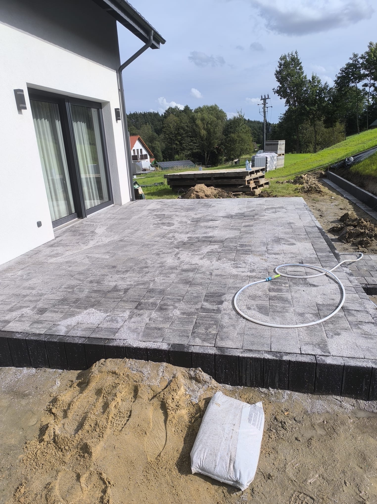 Szara kostka brukowa tworząca taras przy domu, z workiem cementu i wężem ogrodowym. Widok na zielone wzgórza i las w tle.