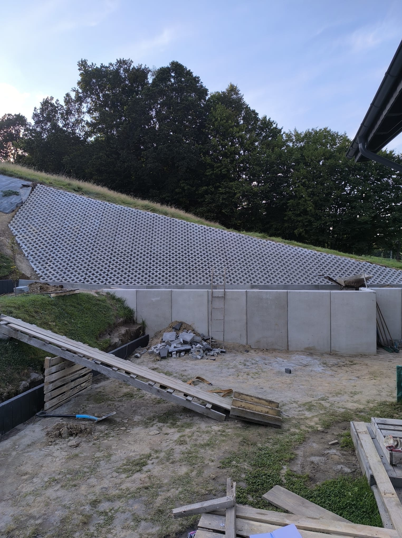 Wzmocnienie skarpy z użyciem ażurowych płyt betonowych i betonowych murków oporowych. Widoczne narzędzia i materiały budowlane. Ujęcie z dołu, w tle drzewa.