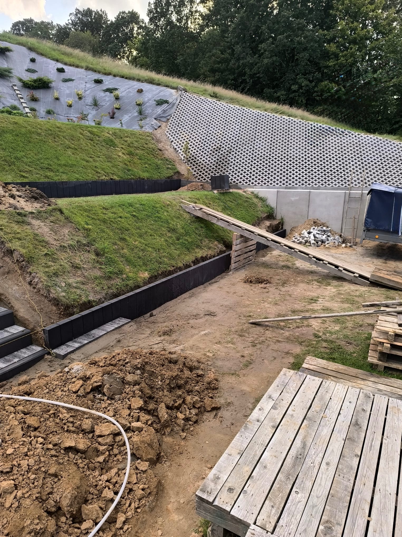 Budowa tarasów w ogrodzie: czarne, betonowe murki oporowe, ziemia, palety. W tle zielony, wzmocniony skarpą teren. Widok na budowę z góry.