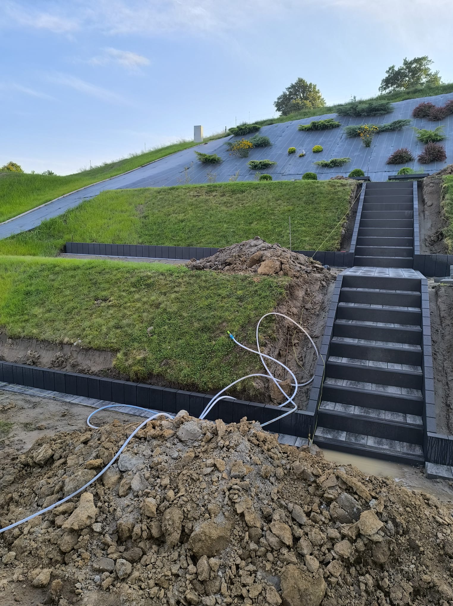 Budowa schodów z kostki brukowej na wzgórzu z roślinami, widoczny etap prac, krawężniki i stos ziemi, biały wąż do podlewania. Nietypowe ujęcie w budownictwie.
