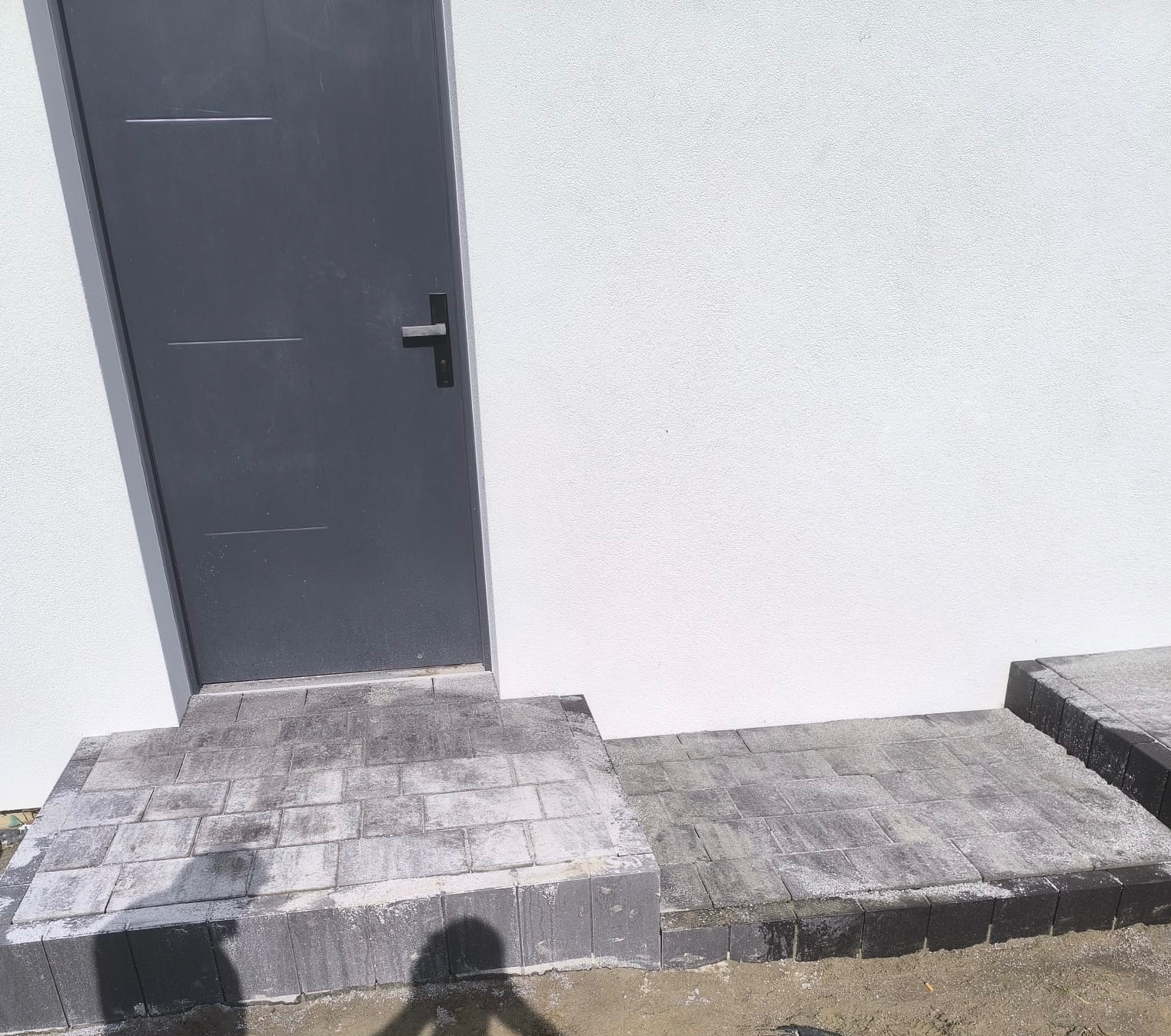 Schody wejściowe z kostki brukowej w odcieniach szarości, prowadzące do szarych drzwi na tle białej ściany budynku. Widoczny fragment powierzchni z kostki.