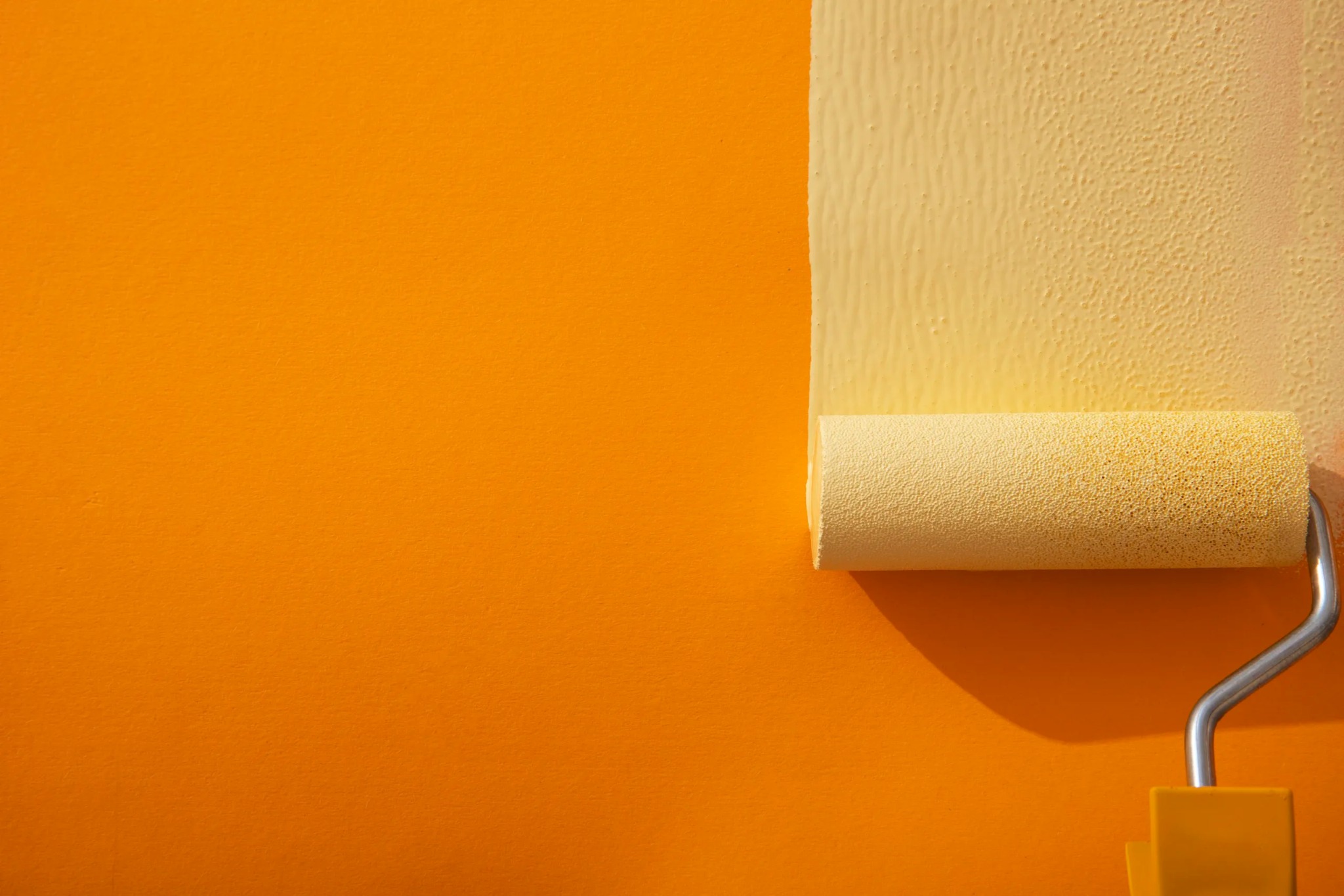 Żółty wałek malarski nanosi farbę na pomarańczową ścianę, tworząc pionowy pas świeżego koloru. Widoczna faktura wałka i farby.