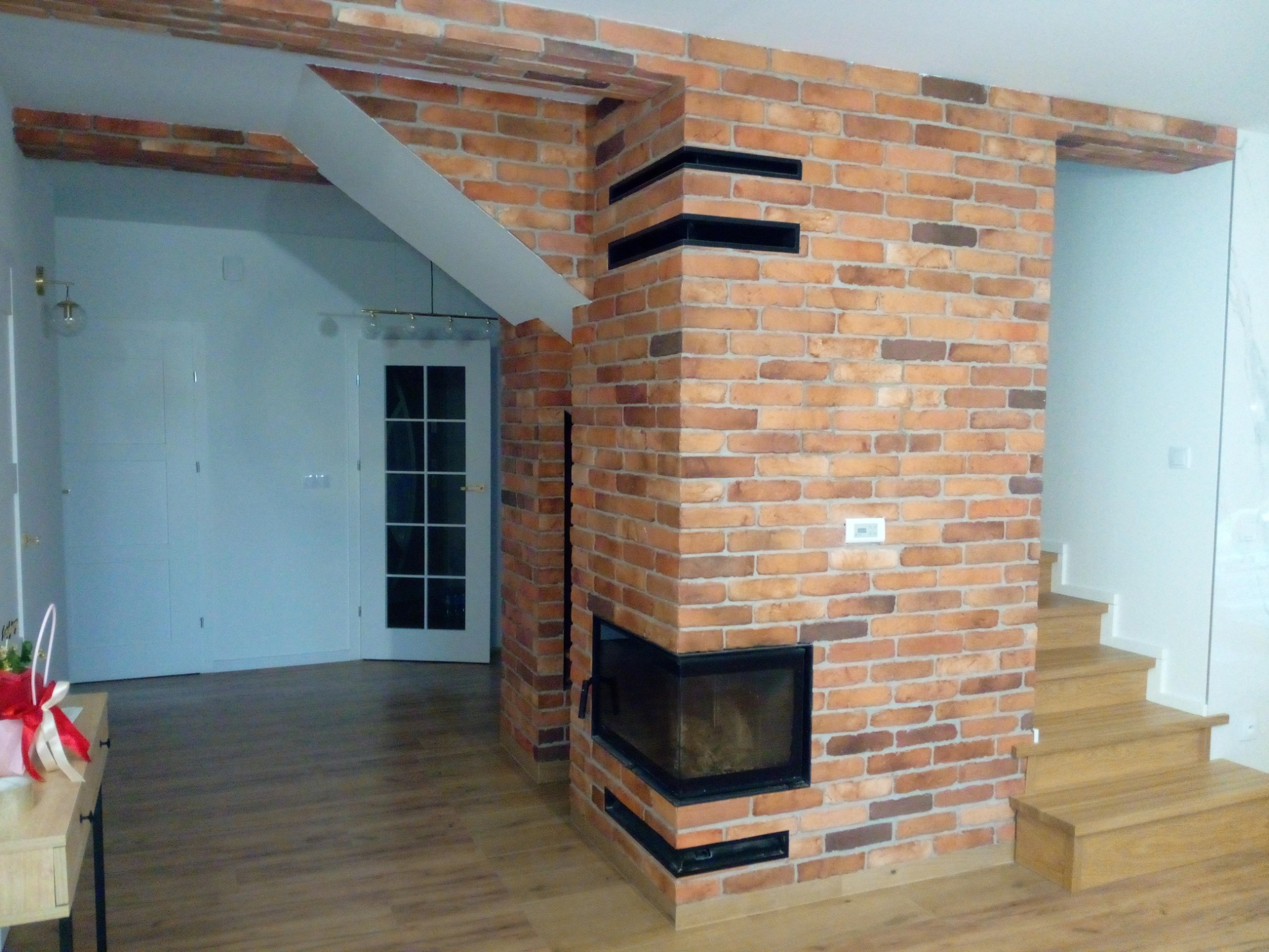 Loftowe wnętrze z ceglaną ścianą i kominkiem narożnym. Widoczne drewniane schody i podłoga. Charakterystyczne, dekoracyjne belki stropowe.