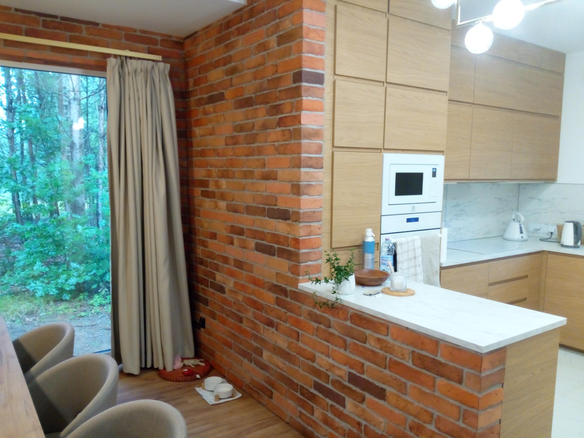 Kuchnia z ceglaną ścianą i jasnymi drewnianymi szafkami. Widok na las przez okno z beżowymi zasłonami. Elementy dekoracyjne na blacie.