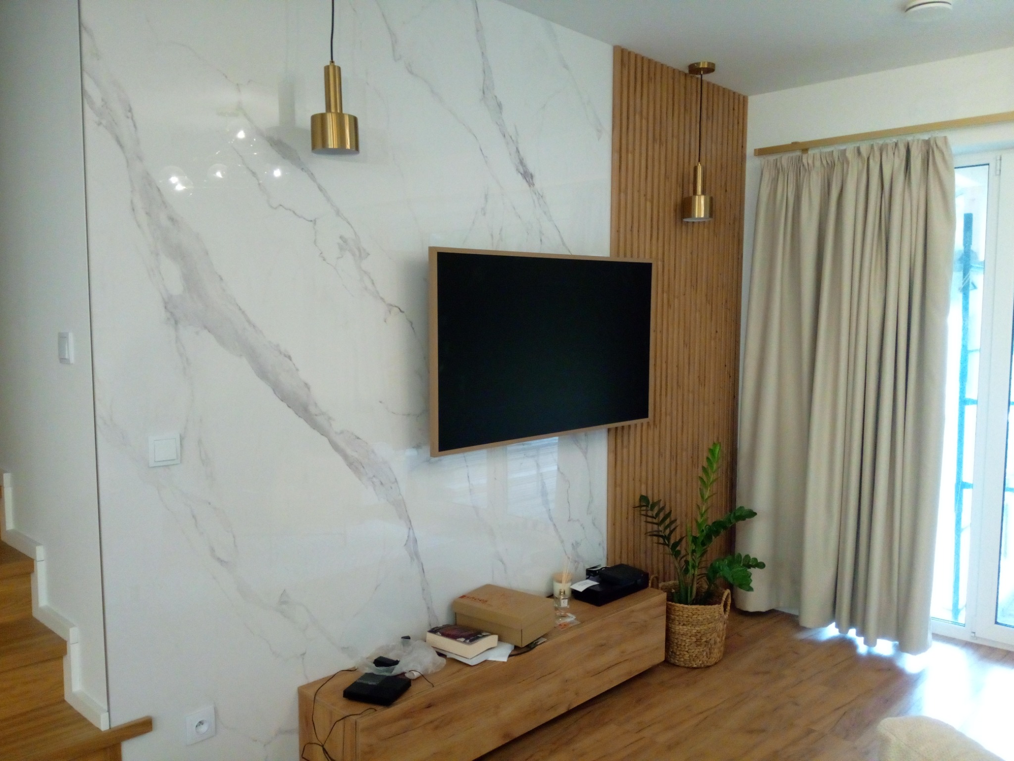 Elegancki salon z marmurową ścianą, drewnianymi lamelami, wiszącym telewizorem i złotymi lampami. Jasne wnętrze z drewnianą podłogą i rośliną doniczkową.