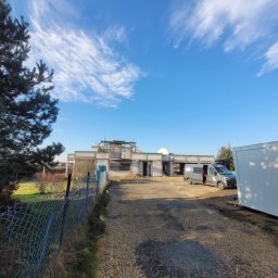 Usb Janek - Surowy stan domu w budowie, z widoczną konstrukcją i szarym vanem na tle błękitnego nieba z chmurami, otoczony zielenią i ogrodzeniem z siatki.