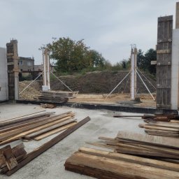 Usb Janek - Wczesny etap budowy domu: surowe ściany z betonu komórkowego, drewniane szalunki wsparte metalowymi stężeniami, deski na posadzce. Widok na plac budowy.