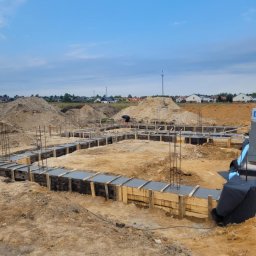Usb Janek - Zalane betonem szalunki fundamentowe z wystającymi prętami zbrojeniowymi na placu budowy. W tle widoczne hałdy ziemi i zabudowa jednorodzinna.