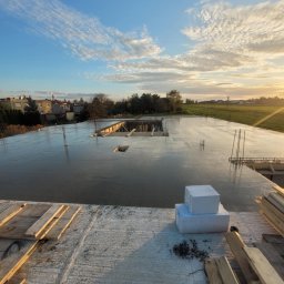 Usb Janek - Zalany strop betonowy domu jednorodzinnego w trakcie budowy, widoczne zbrojenia, szalunki i bloczki styropianowe. W tle krajobraz z zachodzącym słońcem.