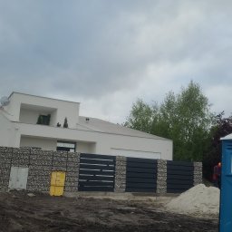 Usb Janek - Nowoczesny dom z gabionowym ogrodzeniem i metalową bramą. Widoczne elementy placu budowy, ziemia i piasek. Minimalistyczna architektura na tle pochmurnego nieba.