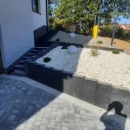 Nowoczesna aranżacja terenu: schody z kostki, rabaty z białym kamieniem, kuliste lampy. Minimalistyczny design, cień fotografa na pierwszym planie.