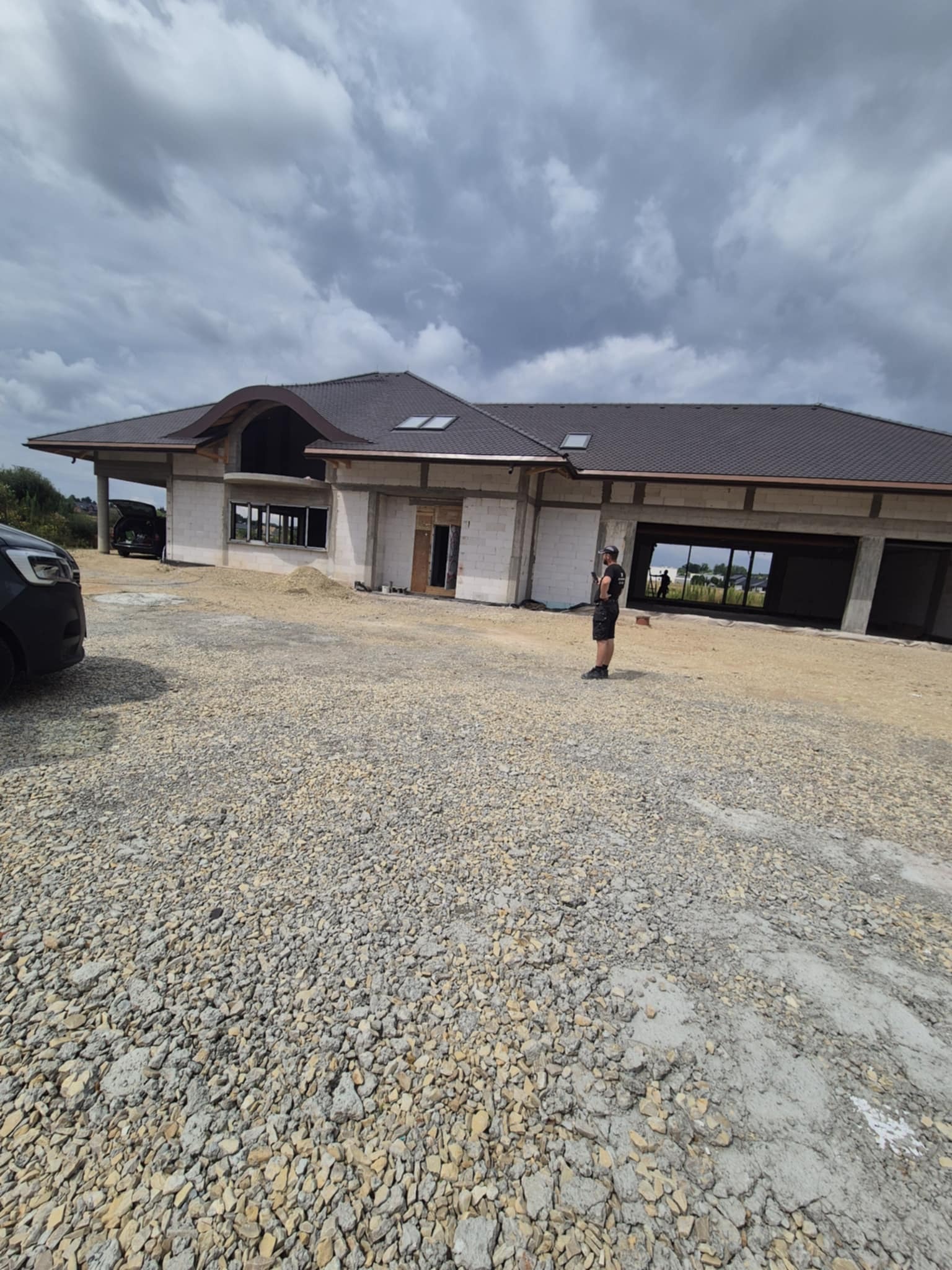 Budowa domu z pustaków: ściany w stanie surowym otwartym, widoczny dach z brązowej dachówki, teren budowy z gruzem, osoba w tle. Dom parterowy z dużymi oknami.