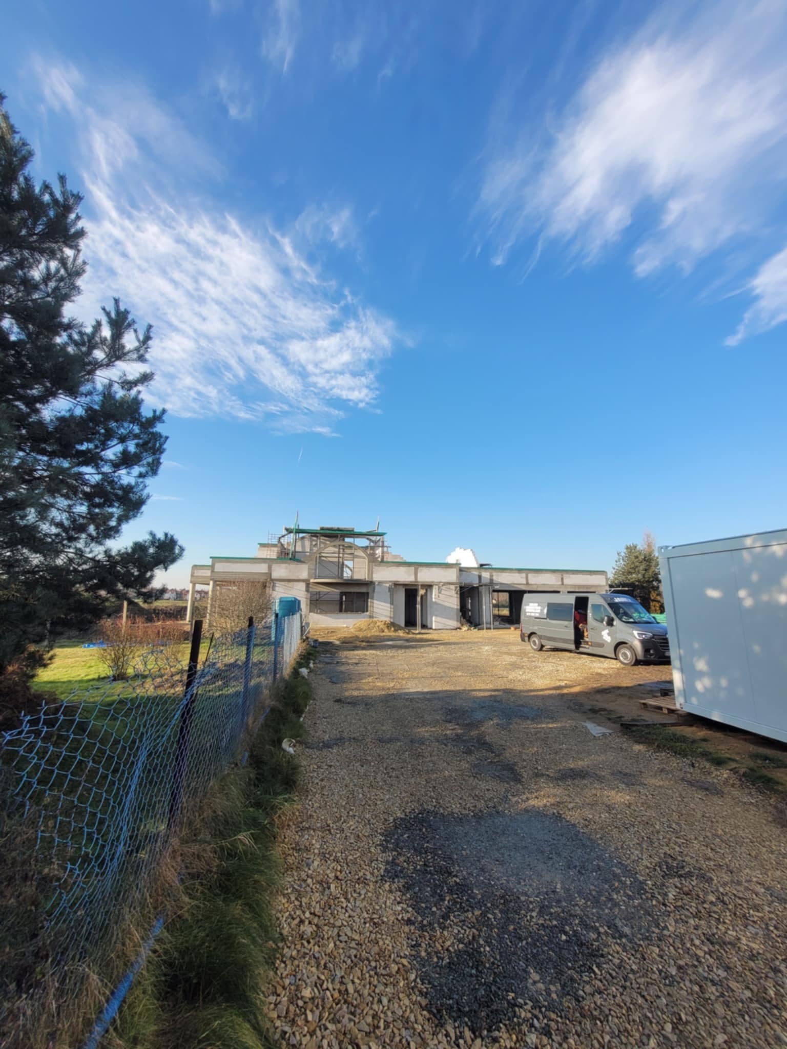 Surowy stan domu w budowie, z widoczną konstrukcją i szarym vanem na tle błękitnego nieba z chmurami, otoczony zielenią i ogrodzeniem z siatki.