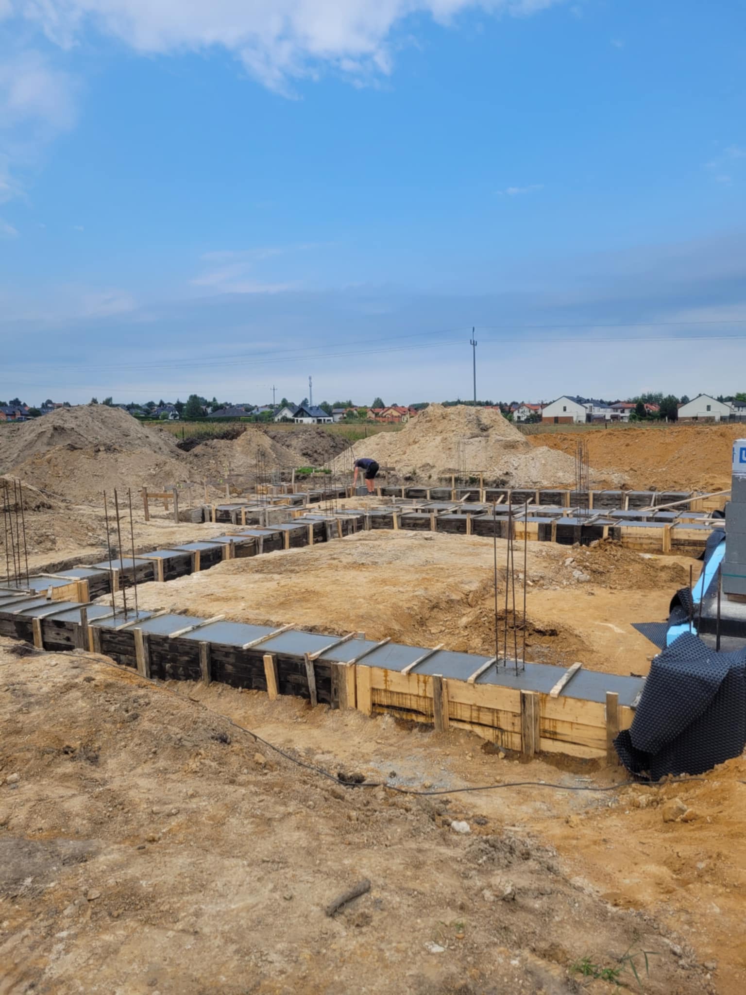Zalane betonem szalunki fundamentowe z wystającymi prętami zbrojeniowymi na placu budowy. W tle widoczne hałdy ziemi i zabudowa jednorodzinna.