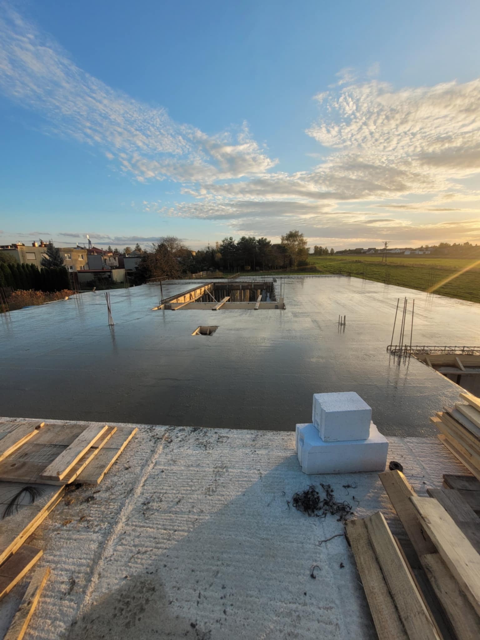 Zalany strop betonowy domu jednorodzinnego w trakcie budowy, widoczne zbrojenia, szalunki i bloczki styropianowe. W tle krajobraz z zachodzącym słońcem.