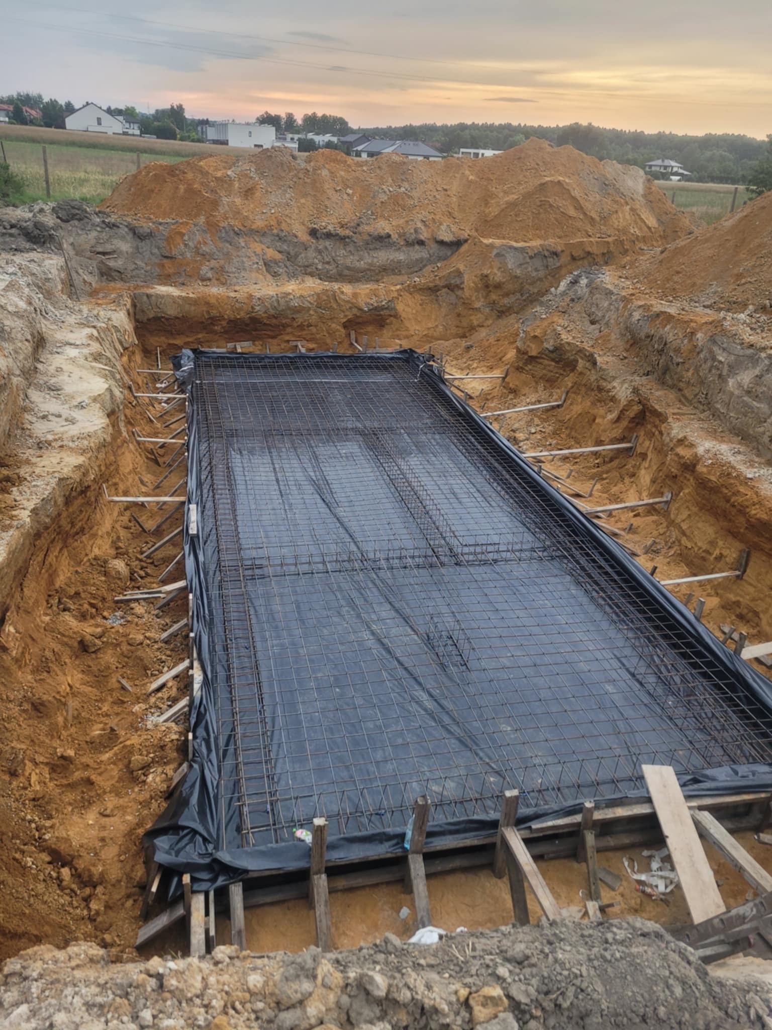 Ziemny wykop z przygotowanym zbrojeniem fundamentów pod budowę domu. Widoczne drewniane szalunki i czarna folia izolacyjna. W tle zabudowa jednorodzinna i niebo o zmierzchu.
