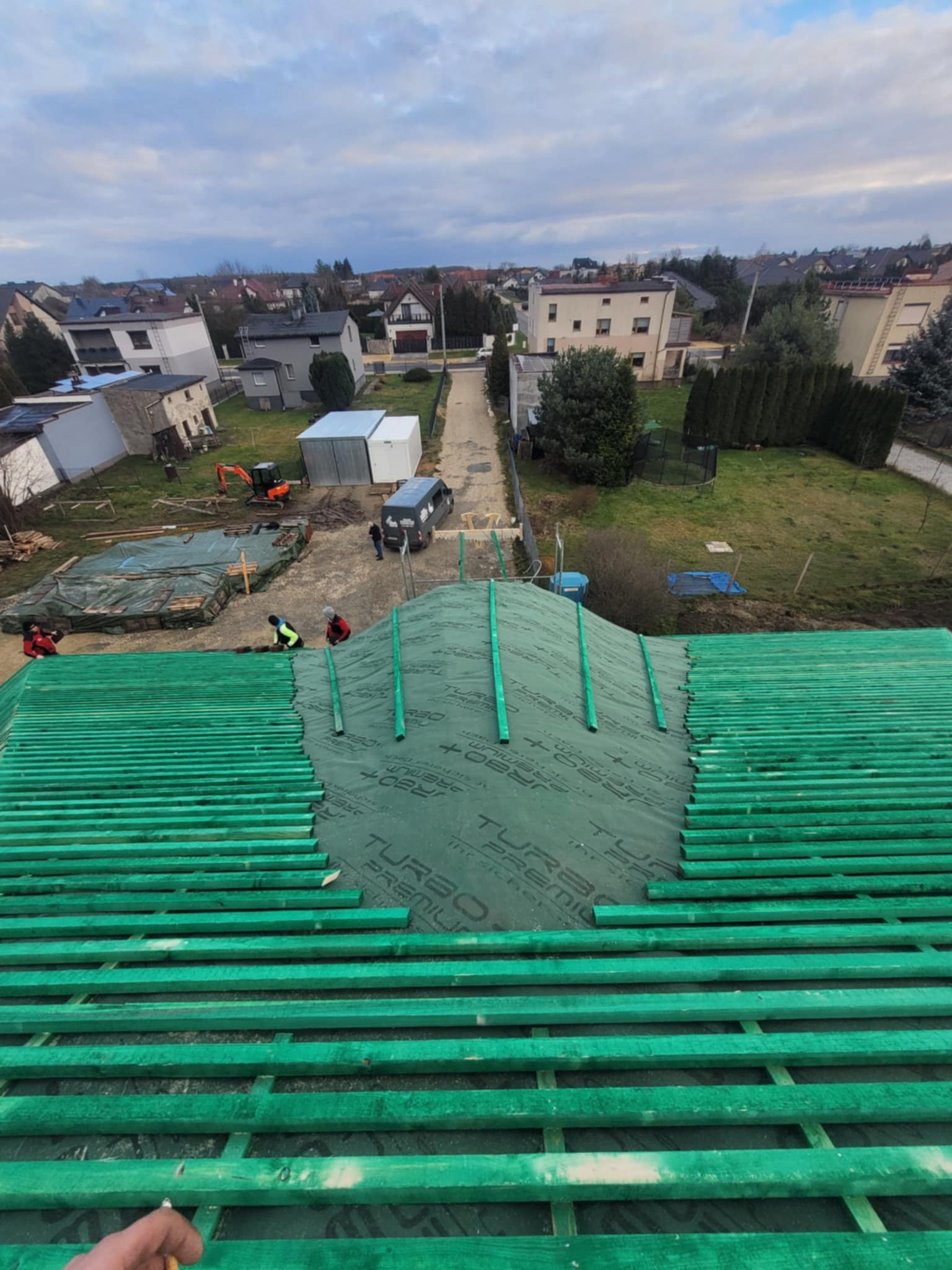 Montaż łat dachowych i membrany Turbo Premium na nowym dachu, widok z góry na tle zabudowy jednorodzinnej i placu budowy z mini koparką.