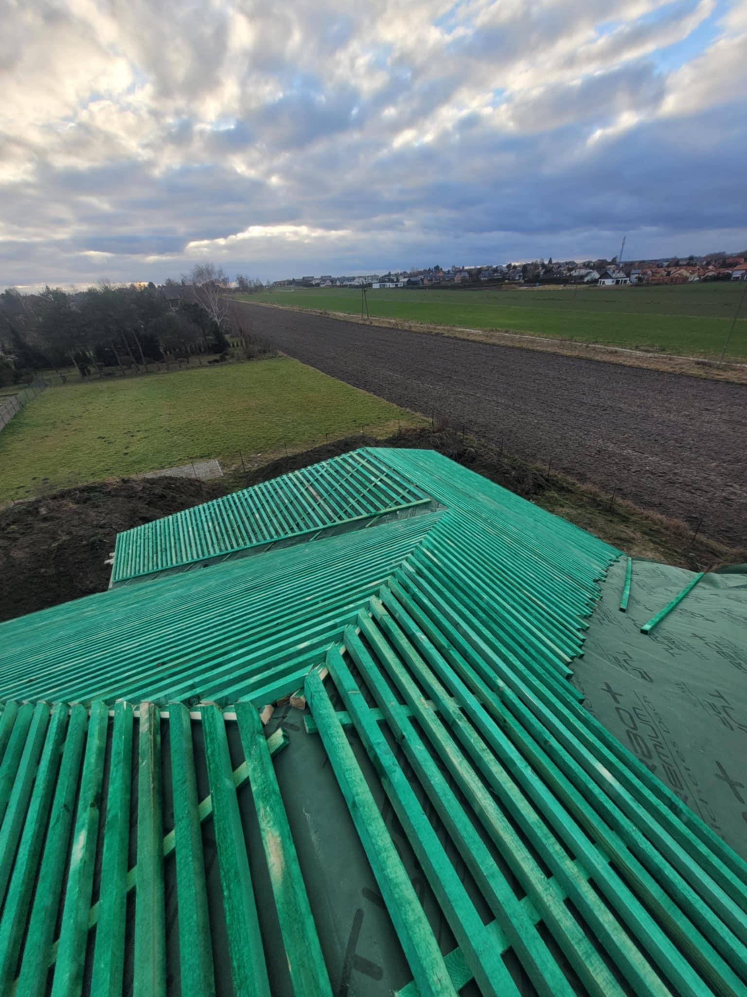 Zielone łaty dachowe na membranie, widok z góry na budowę dachu domu jednorodzinnego. W tle pola uprawne i zabudowa wiejska, pochmurne niebo.