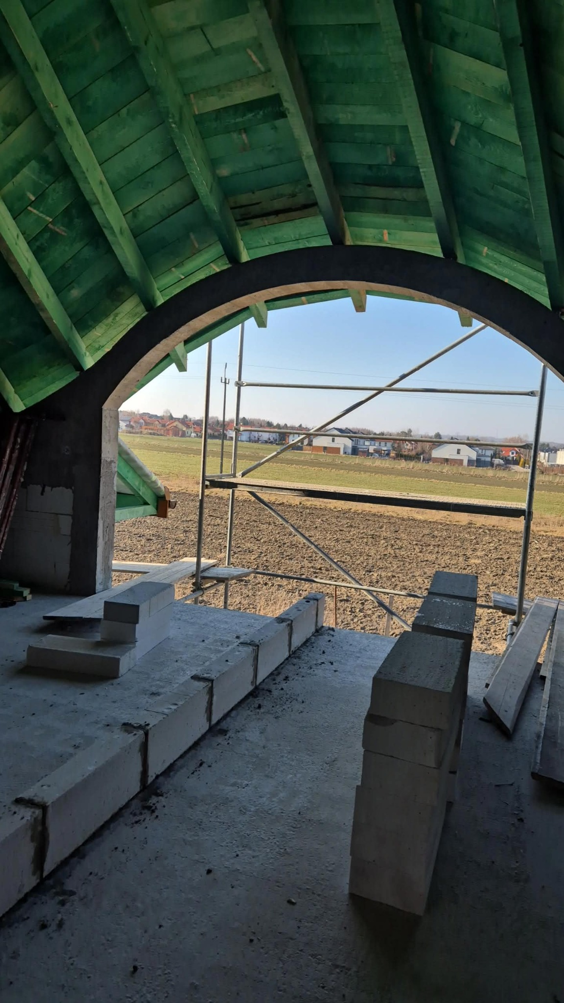 Wnętrze domu w budowie z widokiem na krajobraz przez łukowate okno. Ułożone bloczki z betonu komórkowego, zielona konstrukcja dachu i rusztowanie.