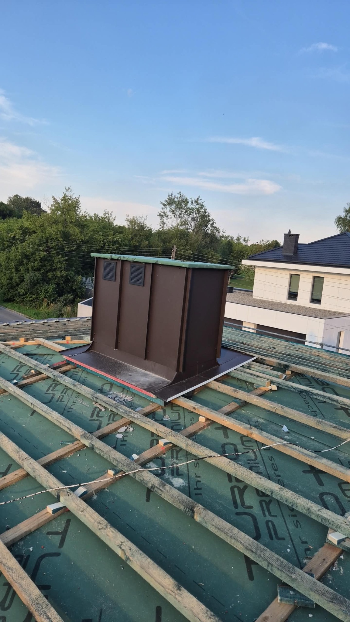 Montaż brązowego komina wentylacyjnego na dachu z zieloną membraną i drewnianą konstrukcją. W tle dom z czarnym dachem i jasną elewacją.