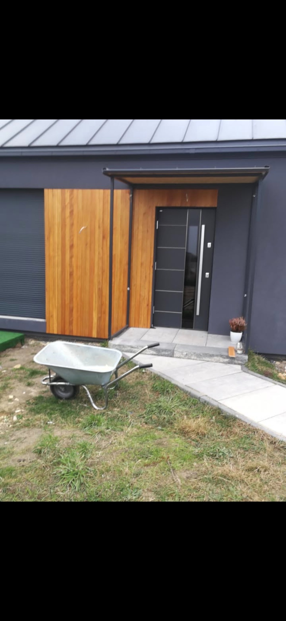 Nowoczesny dom z szarą elewacją, drewnianym panelem i wózkiem budowlanym na trawniku. Minimalistyczny styl, widoczne detale wykończenia.