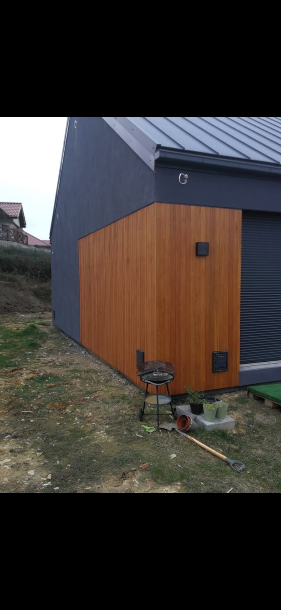 Elewacja budynku z szarego tynku i drewnianej okładziny. Widoczny grill, łopata i doniczki z roślinami. Ujęcie z dołu, na zewnątrz.