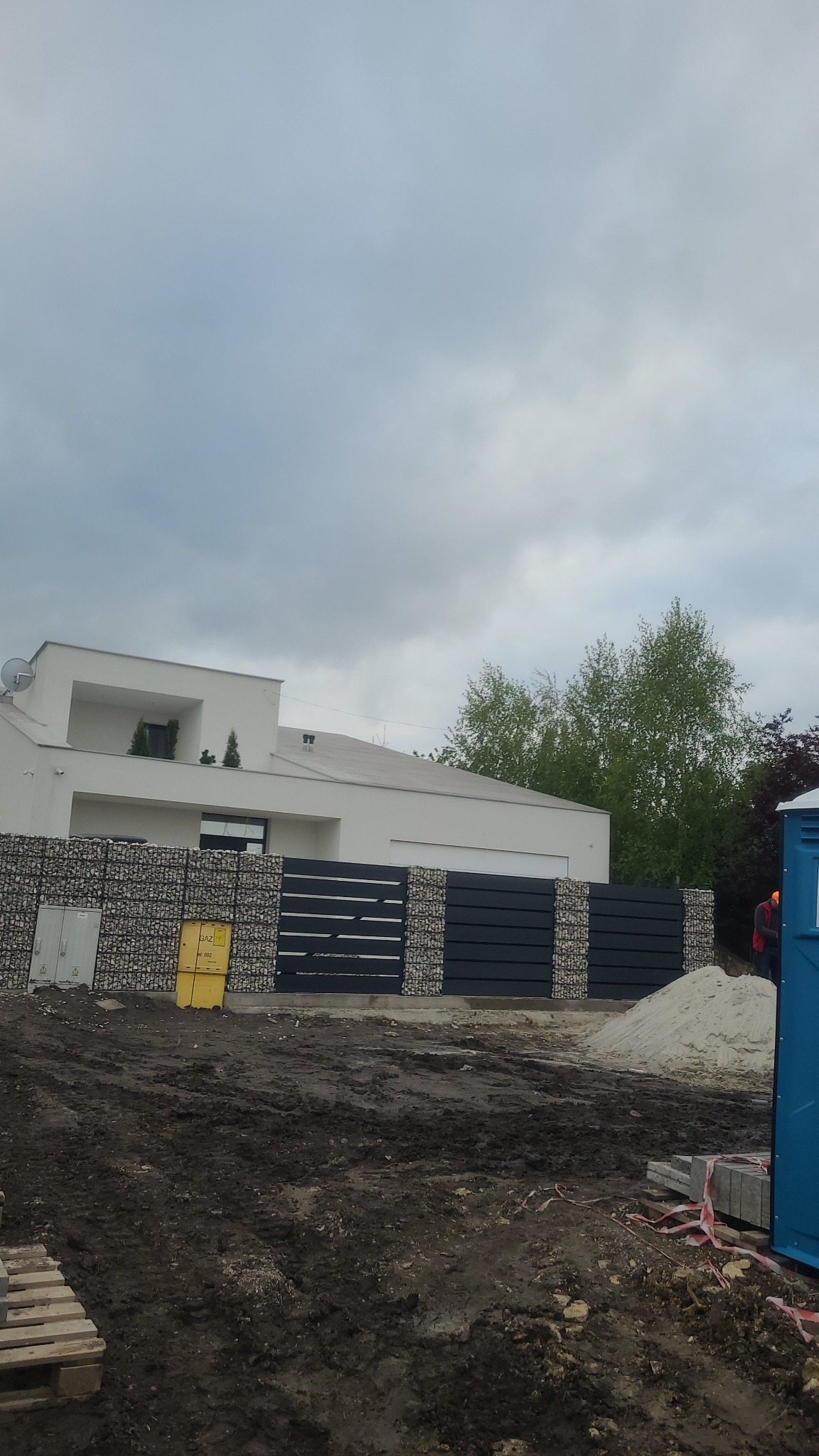 Nowoczesny dom z gabionowym ogrodzeniem i metalową bramą. Widoczne elementy placu budowy, ziemia i piasek. Minimalistyczna architektura na tle pochmurnego nieba.