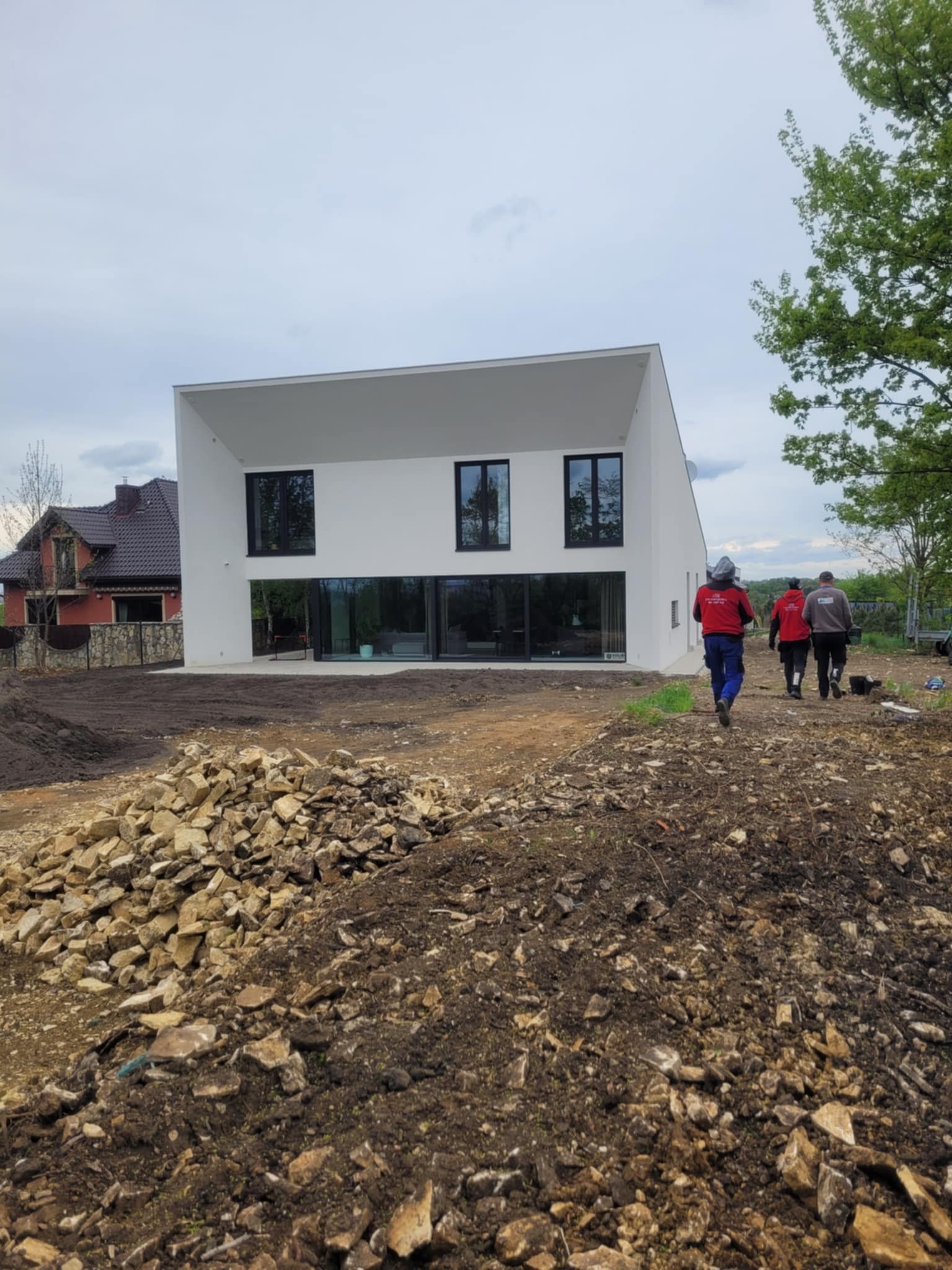 Nowoczesny, biały dom z płaskim dachem i dużymi oknami, widziany z perspektywy placu budowy. Trzy osoby oddalają się od budynku. W tle drugi dom.