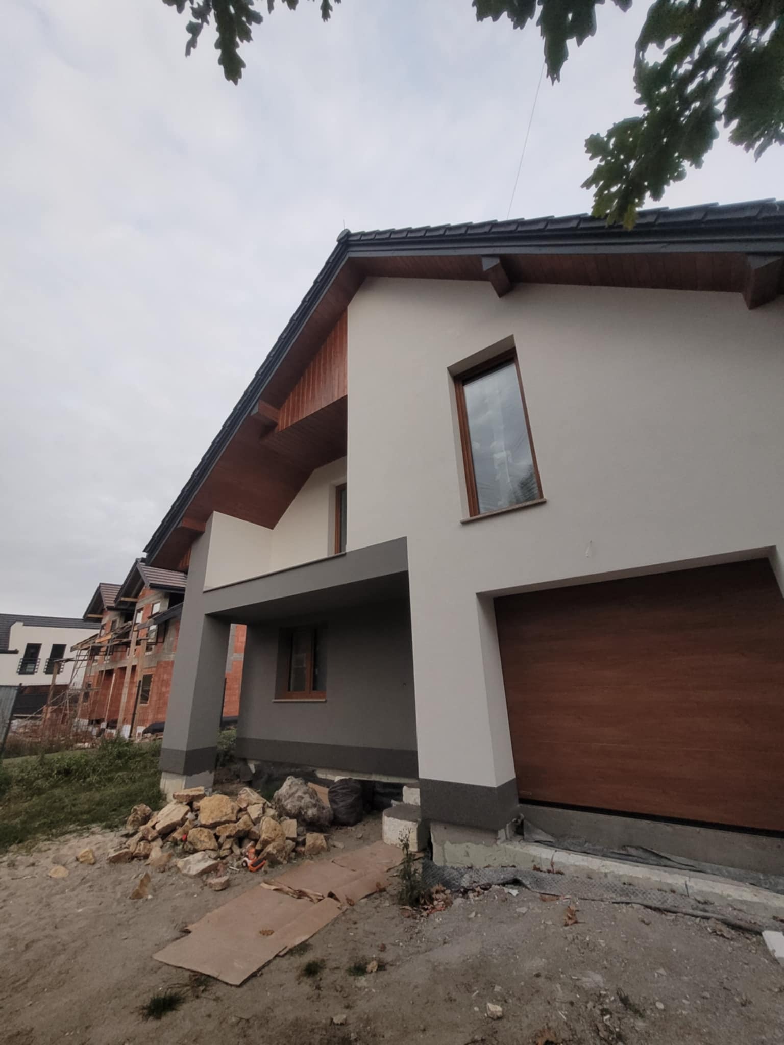 Nowoczesny dom jednorodzinny z garażem w Wilkowisku, świeżo po wykonaniu elewacji. Widoczne detale wykończeniowe i sąsiednie budynki w trakcie budowy.