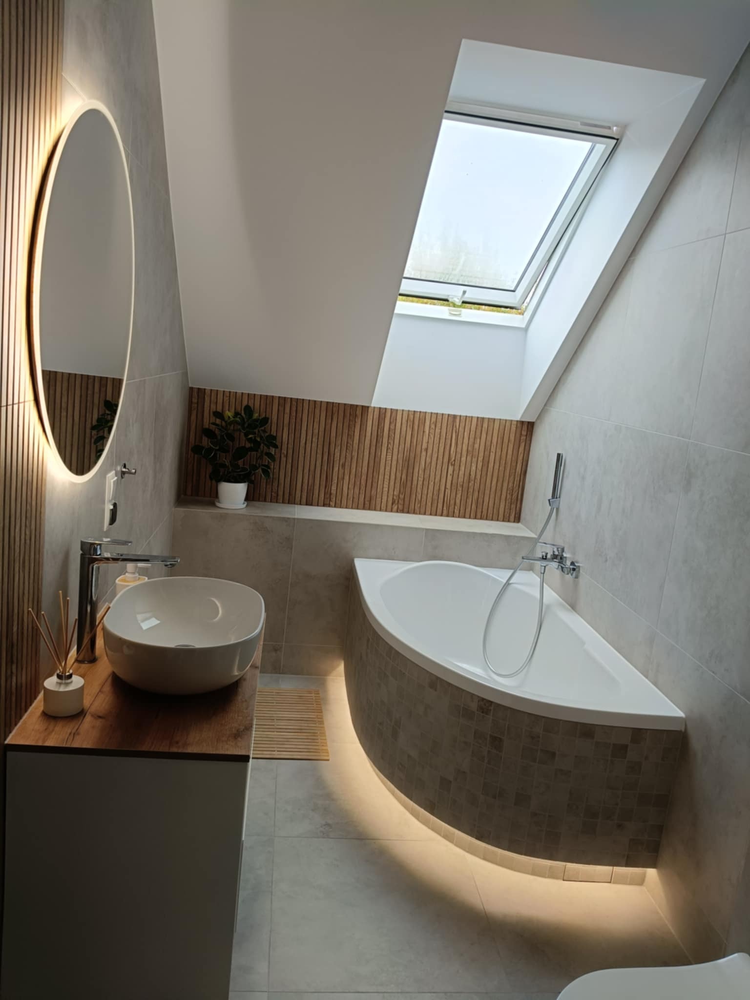 Nowoczesna łazienka na poddaszu z owalnym lustrem LED, skośnym oknem dachowym i podświetloną wanną narożną. Umywalka nablatowa na drewnianym blacie, szare płytki.