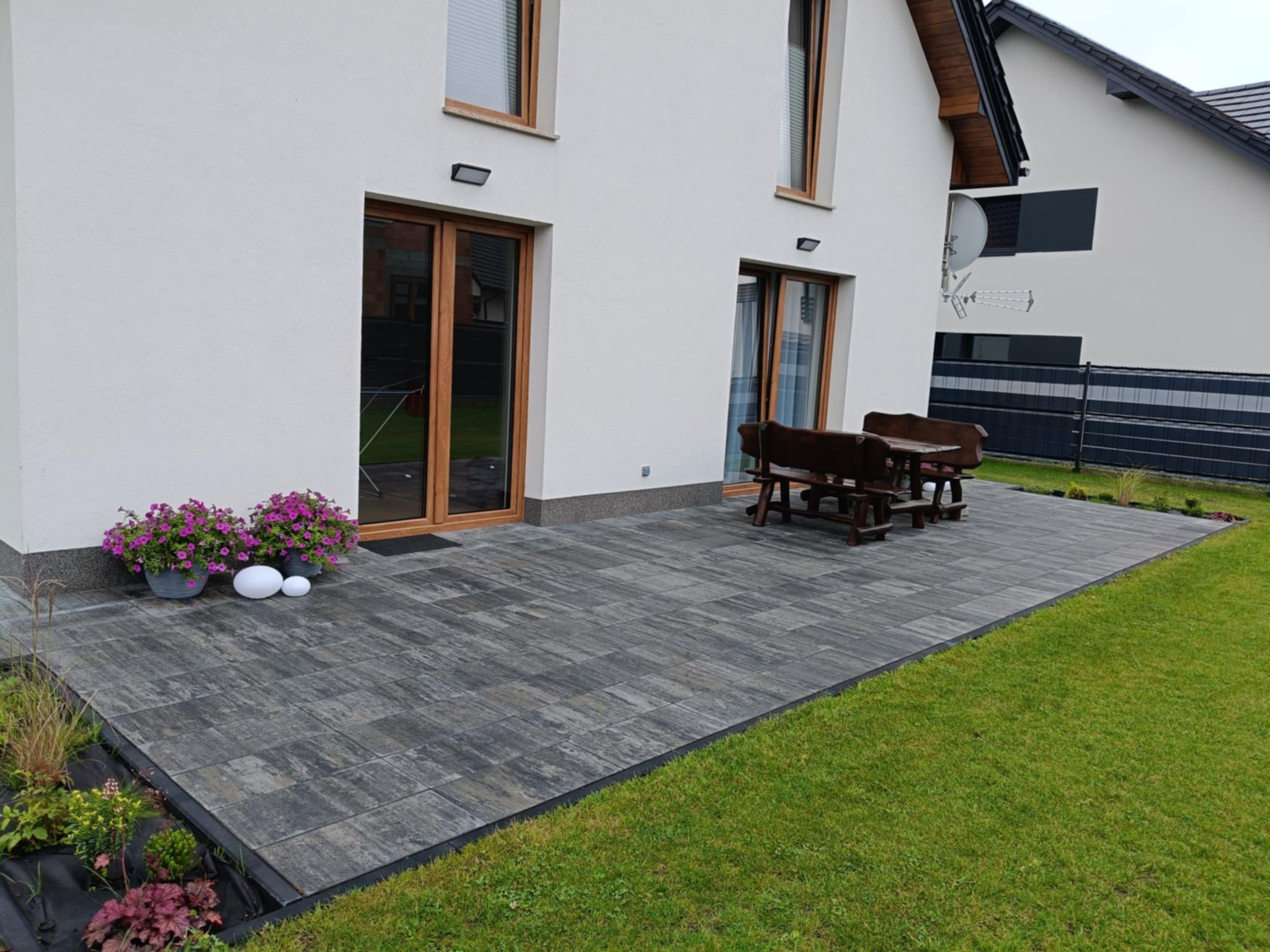 Nowoczesny taras z szarej kostki brukowej przy białym domu z drewnianą stolarką okienną. Uwagę przyciąga rabata z roślinami oddzielająca taras od trawnika.